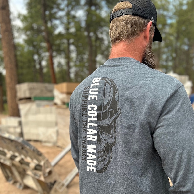 May include: Dark gray long-sleeve shirt with a skull graphic wearing a hard hat. The text "BLUE COLLAR MADE SUPPORT THE TRADES" is printed vertically on the back of the shirt in white. The shirt is made of a soft material.