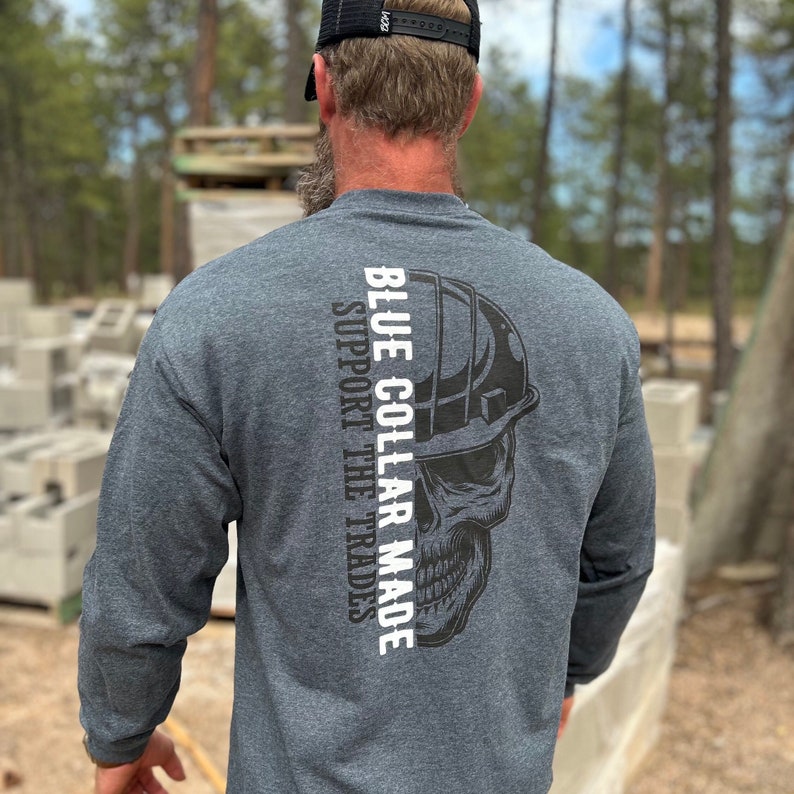 May include: Dark gray long-sleeve shirt with the text "BLUE COLLAR MADE SUPPORT THE TRADES" in white. A black skull graphic wearing a hard hat is printed on the back. The shirt is made of a soft, comfortable material.