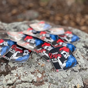Pu&ograve; includere: Un mucchio di adesivi in ​​bianco e nero con un design Rosie la Rivettatrice rosso, bianco e blu. L'adesivo presenta uno scheletro con una bandana rossa e il testo "Support Rosies in the Trades".