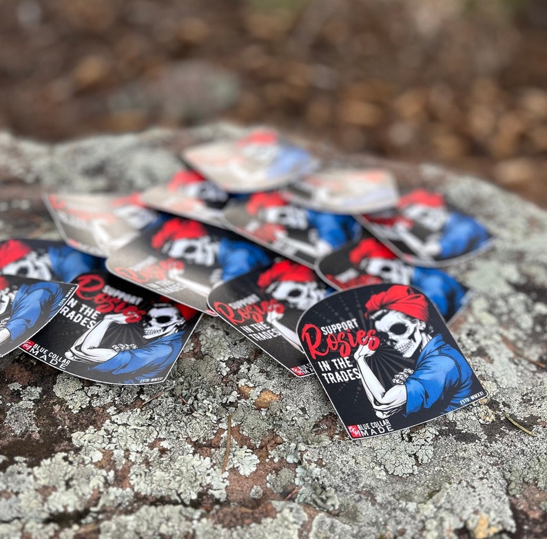 Pu&ograve; includere: Un mucchio di adesivi in ​​bianco e nero con un design Rosie la Rivettatrice rosso, bianco e blu. L'adesivo presenta uno scheletro con una bandana rossa e il testo "Support Rosies in the Trades".