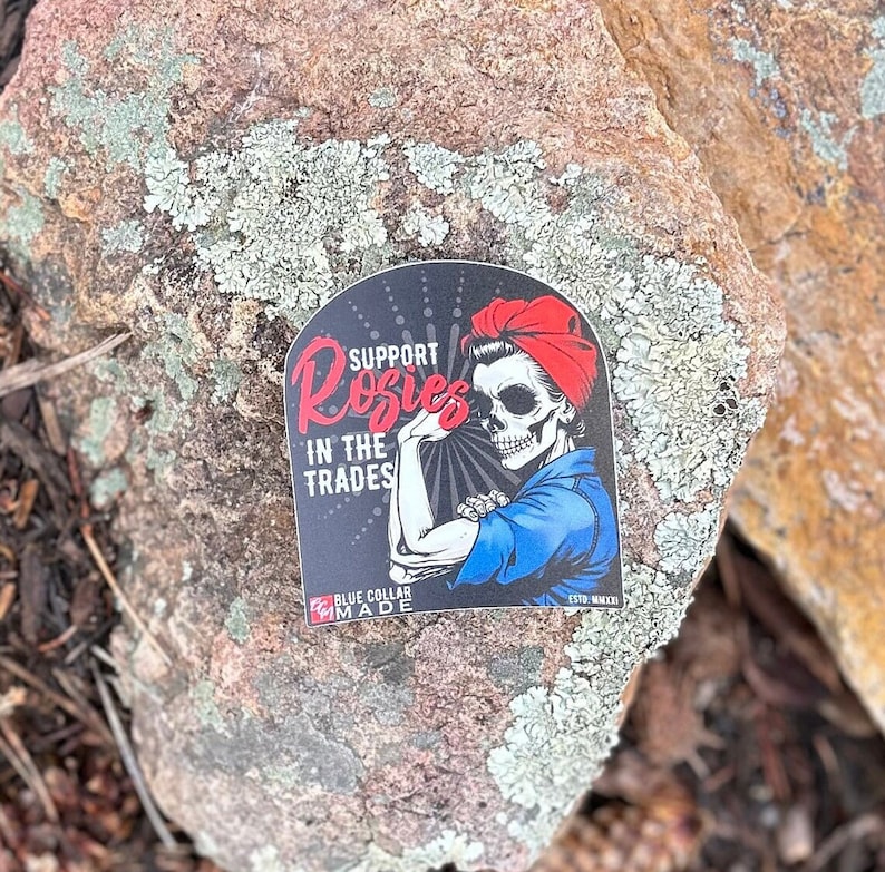 Pu&ograve; includere: Un adesivo nero e blu con un'immagine di cartone animato di una donna con un viso di teschio che indossa un bandana rosso e una camicia blu. Il testo sull'adesivo recita "Support Rosies in the Trades" e "Blue Collar Made".