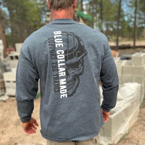 May include: A dark gray long-sleeve shirt with the text "BLUE COLLAR MADE SUPPORT THE TRADES" printed vertically on the back. A black and white skull graphic wearing a hard hat is next to the text. The shirt is made of a soft material.