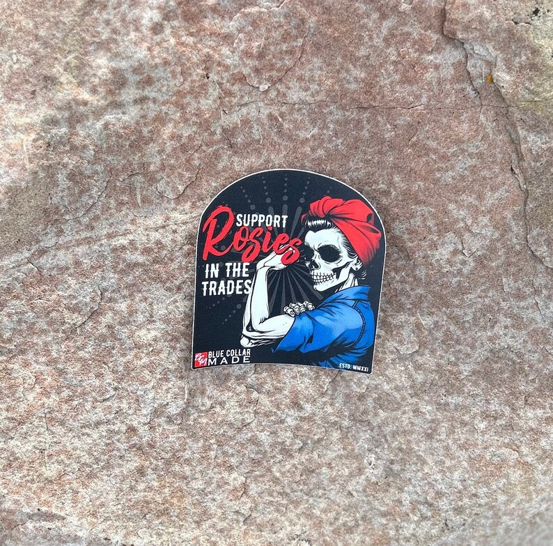 Pu&ograve; includere: Un adesivo in bianco e nero con una donna con un viso di teschio che indossa una camicia blu e una bandana rossa. L'adesivo dice "Support Rosies in the Trades" e "Blue Collar Made".