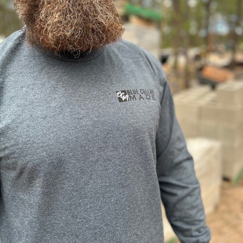 May include: A gray long-sleeve shirt with the text "BLUE COLLAR MADE" in black. The shirt is made of a soft material and has a crew neck. The image is taken outdoors.