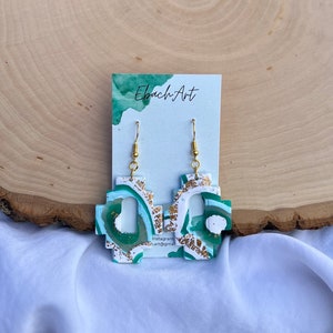 May include: A pair of green and white geometric earrings with gold accents. The earrings are made of polymer clay and have a unique, abstract design. The earrings are hanging from gold hooks.