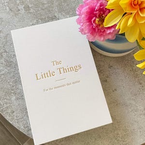 May include: A white rectangular book with gold embossed text that reads The Little Things For the moments that matter. The cover features a clean, matte finish, and the book is positioned next to a vibrant bouquet of pink and yellow flowers.