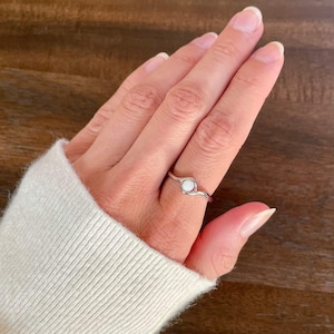 May include: A silver-toned ring featuring a small, round, white opal-like stone set within a delicate, wavy metal band. The ring is worn on a finger, showcasing the polished finish of the metal and the subtle iridescent glow of the central stone.