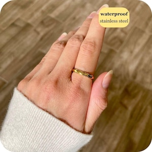 May include: A gold-colored ring with a textured, faceted surface. The ring is made of stainless steel and is labeled as waterproof. The band features a repeating geometric pattern that reflects light, creating a subtle shimmer across the metal surface.