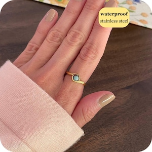 May include: A gold-toned stainless steel ring featuring a small, round, light blue stone set in the center. The band has a smooth, curved design that wraps around the stone. The text waterproof stainless steel is visible in the image.