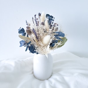 May include: A white ceramic vase filled with a dried flower arrangement featuring white and blue dried flowers. The bouquet includes dried lavender, white bunny tails, and eucalyptus.