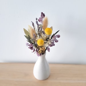 May include: A small white vase containing a bouquet of dried flowers. The flowers are a mix of purple, yellow, and white. The vase is sitting on a light brown wooden surface.