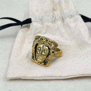 May include: A gold-colored ring featuring a detailed face design. The ring has a rectangular face with a sculpted face and curly hair. The ring is resting on a white cloth bag with a black drawstring.
