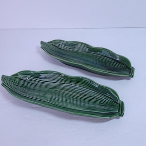 Puede incluir: Dos fuentes de cerámica verde con forma de hojas de maíz. Las fuentes tienen una superficie texturizada y acanalada y un acabado brillante. Las fuentes son de un color verde intenso y rico y son ideales para servir aperitivos o snacks.
