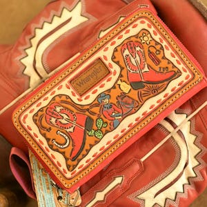 May include: A red leather wallet with a Western-themed design featuring a cowboy, boots, and a horseshoe, resting on a pair of red cowboy boots. The wallet is branded with the Wrangler logo.