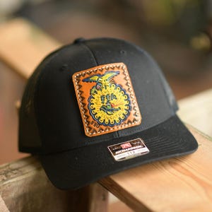 May include: Black baseball cap with a leather patch featuring a colorful emblem. The patch has a yellow and blue design with the letters "FFA". The cap is sitting on a wooden surface.