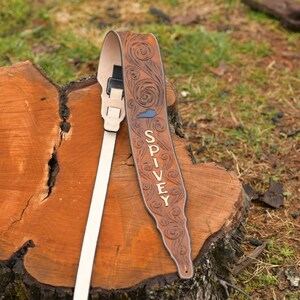 May include: A brown leather guitar strap with a floral design and the name "SPIVEY" embossed in white. The strap has a white adjustable strap and a black buckle.