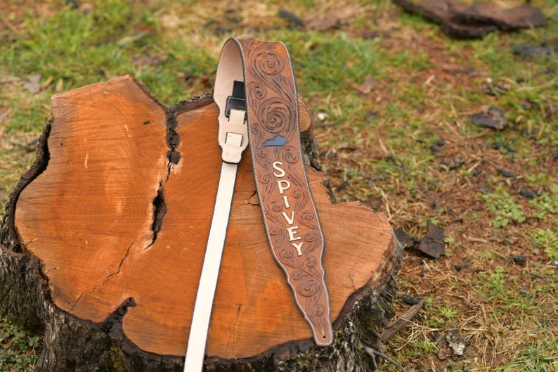 May include: A brown leather guitar strap with a floral design and the name "SPIVEY" embossed in white. The strap has a white adjustable strap and a black buckle.
