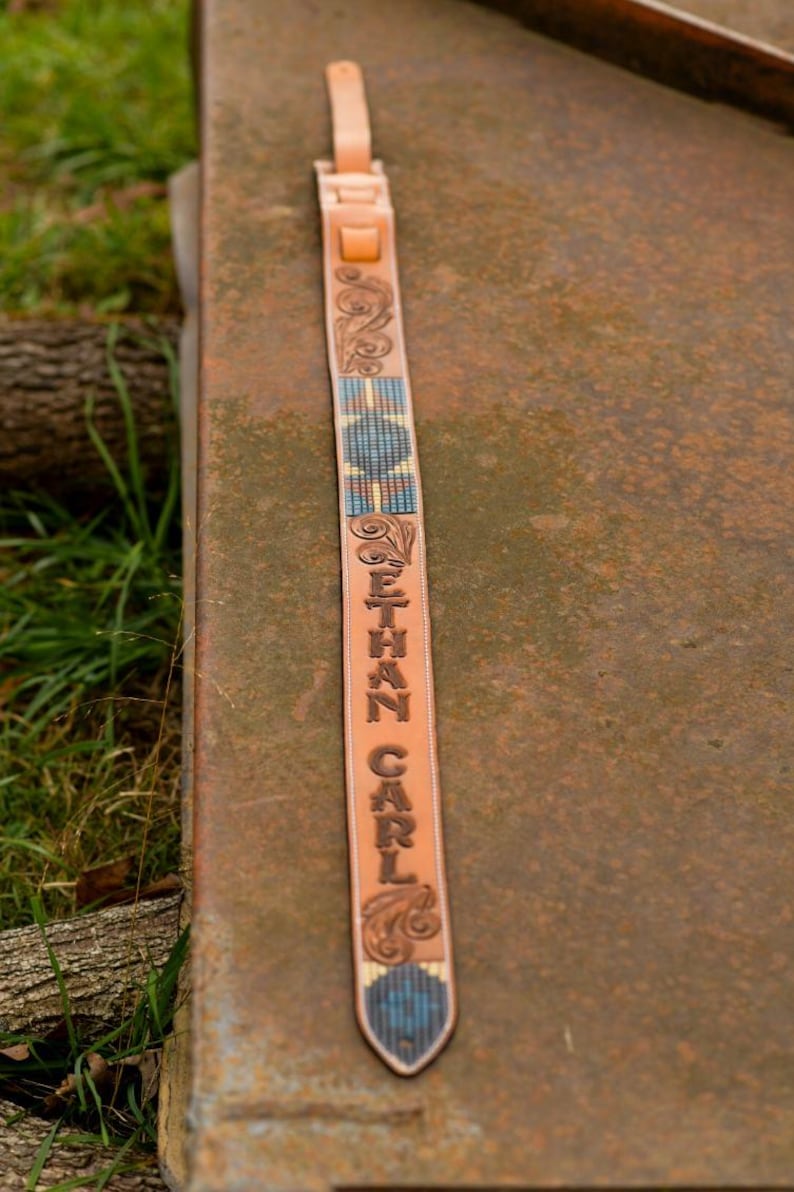May include: A brown leather guitar strap with a blue and white beaded design. The strap is personalised with the name "ETHAN CARL" in a decorative font.