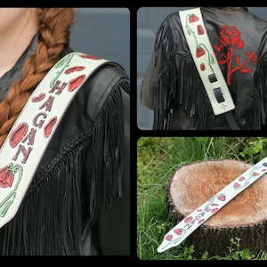 May include: White leather guitar strap with red and green floral designs and the name "HAGAN" written in black.