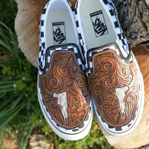 May include: A pair of slip-on shoes with a brown leather design featuring a steer skull and floral accents. The shoes have a white sole and a black and white checkered pattern on the sides.