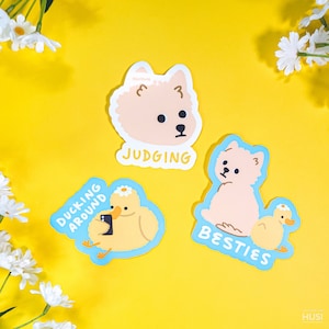 Könnte beinhalten: Drei gestanzte Aufkleber auf gelbem Hintergrund. Einer zeigt einen hellbraunen Hund mit dem Wort "JUDGING". Ein anderer zeigt eine gelbe Ente, die ein Foto mit dem Text "DUCKING AROUND" macht. Der letzte Aufkleber hat einen hellbraunen Hund und ein Entlein mit dem Wort "BESTIES".