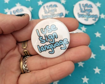 May include: A white button with blue text that reads "I use sign language". The button is being held in a hand.