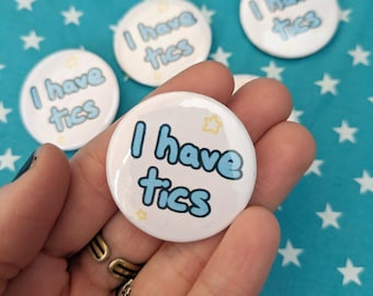 May include: Four white round buttons with blue text that reads "I have tics" and yellow stars. The buttons are held in a hand.