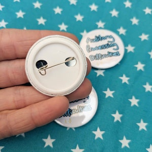 May include: The back of a white button with a silver pin. The front of the button is visible in the background and says "Auditory Processing Difficulties".