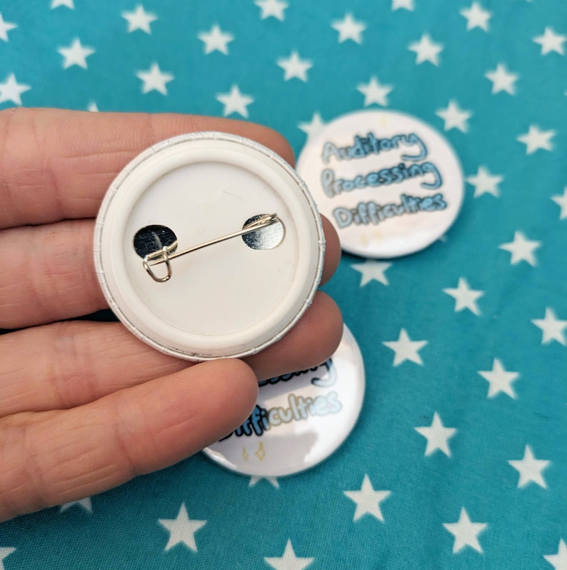 May include: The back of a white button with a silver pin. The front of the button is visible in the background and says "Auditory Processing Difficulties".