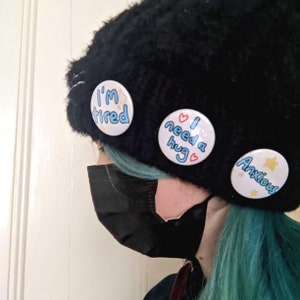 May include: A person wearing a black beanie with three buttons pinned to it. The buttons say "I'm tired", "I need a hug", and "Anxious". The person is also wearing a black face mask and has teal blue hair.