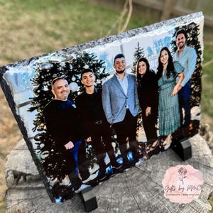May include: A family portrait printed on a black slate coaster. The photo shows five people, including two men, two women, and a young boy, standing in front of a Christmas tree. The coaster has a rough, textured surface.