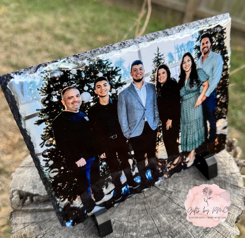 May include: A family portrait printed on a black slate coaster. The photo shows five people, including two men, two women, and a young boy, standing in front of a Christmas tree. The coaster has a rough, textured surface.