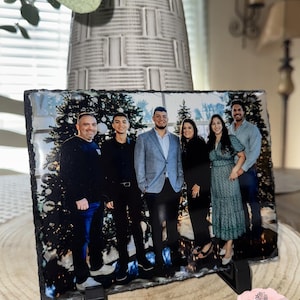 May include: A black slate photo holder with a printed image of a family of six standing in front of a Christmas tree. The family is dressed in casual clothing and smiling. The photo holder has a black stand.