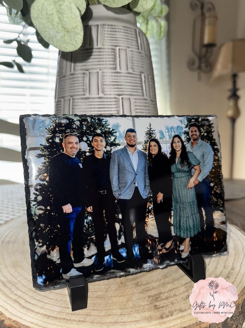 May include: A black slate photo holder with a printed image of a family of six standing in front of a Christmas tree. The family is dressed in casual clothing and smiling. The photo holder has a black stand.