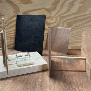 May include: A wooden mousetrap with a metal spring and a wooden base. The mousetrap is surrounded by four pieces of wood, two of which are painted black.