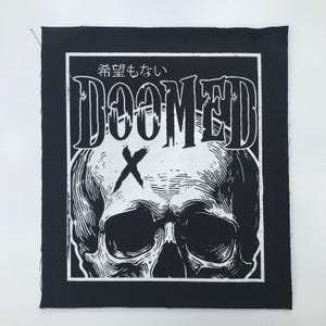 May include: Black and white patch featuring a skull graphic with the word "DOOMED" in bold letters. Above the word "DOOMED" is Japanese text. A white border frames the design on a black background.