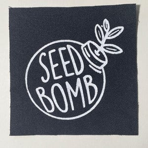 May include: A black square patch with a white graphic of a seed bomb. The design features the words "SEED BOMB" inside a circle, with a plant sprouting from the top. The patch is made of a textured fabric.