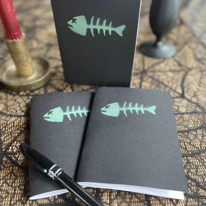 Könnte beinhalten: Kleine schwarze Notizbücher mit einem grünen Fischskelett-Design. Ein schwarzer Stift liegt vor den Notizbüchern. Eine rote Kerze und ein schwarzes Dekorationsobjekt befinden sich im Hintergrund.