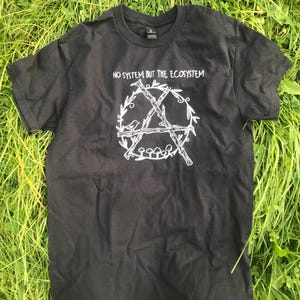May include: Black t-shirt with a white graphic of a stylized anarchy symbol with a bird, branches, and mushrooms inside a wreath. The text "NO SYSTEM BUT THE ECOSYSTEM" is printed above the graphic.