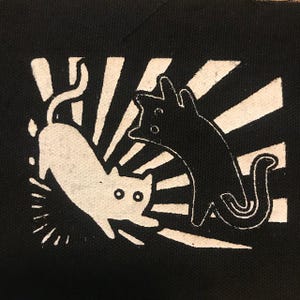 May include: Black and white illustration of two cats on a black background. The cats are facing opposite directions and are surrounded by a white sunburst pattern.