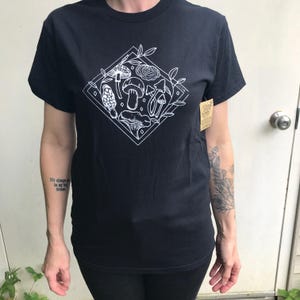May include: Black t-shirt featuring a white diamond-shaped graphic with various mushroom illustrations and leafy accents. The shirt has a crew neck and short sleeves. A tag is attached to the shirt. The overall theme is nature and botanical.