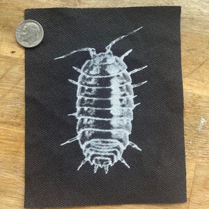 May include: A black fabric patch featuring a detailed white illustration of a woodlouse. The woodlouse is depicted with segmented body and multiple legs. A dime is placed next to the patch for size reference.