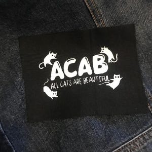 May include: Black patch with white text that reads "ACAB" and "All cats are beautiful". The patch features several stylized white cat silhouettes.