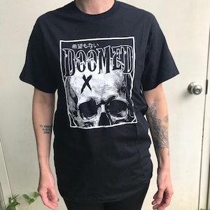 May include: Black t-shirt featuring a white graphic of a skull with the word "DOOMED" above it and the Japanese text "希望もない" above the word "DOOMED". The graphic also includes a white "X" over the skull.