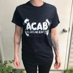 May include: Black t-shirt with white text and cat illustrations. The text reads "ACAB ALL CATS ARE BEAUTIFUL". The shirt has a crew neck and short sleeves.