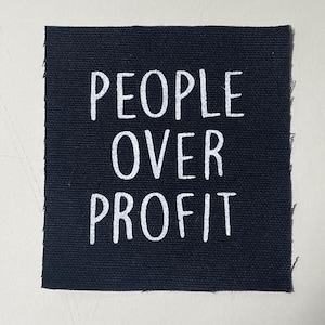 May include: A square, black fabric patch with the words "PEOPLE OVER PROFIT" printed in white, sans-serif font. The patch has a slightly frayed edge and is set against a neutral background.
