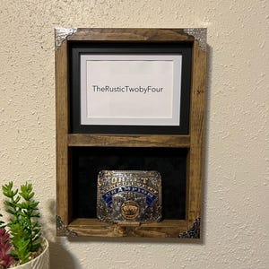May include: A rustic, wooden display case with a black interior. The case features a framed area with the text "TheRusticTwobyFour" and a lower compartment holding a championship belt. Decorative metal corner accents add detail.