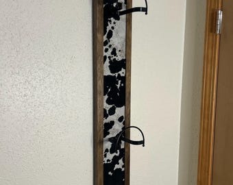May include: A rustic wall-mounted hat rack with a cowhide pattern. The rectangular frame is made of wood, and the center features a black and white cowhide design. Two black metal hat holders are attached to the frame, providing a stylish storage solution.