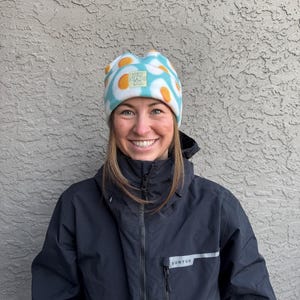 May include: A woman wearing a black jacket with a white and yellow egg-patterned beanie. The jacket has a zipper and a silver zipper pull with the word "BURTON" on it.
