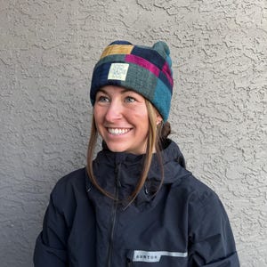 May include: A woman wearing a black jacket with the word "BURTON" on the front. She is wearing a multi-colored plaid beanie with a small logo that says "NO BAD DAYS CO."